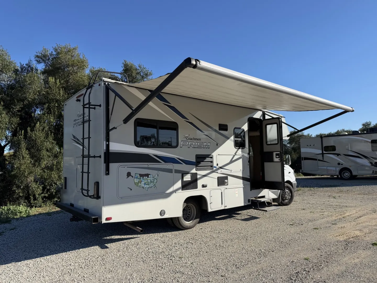 2020 Coachmen Prism 2150CB Class C motorhome with its awning out, parked on gravel under oak trees